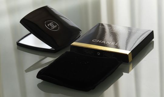 シャネルのコンパクトミラー、ベロア袋、箱がテーブルに置かれたシンプルでエレガントな商品紹介写真。高級感溢れるパッケージが贈り物に最適。