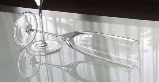 ティファニー商品一覧ページ：テーブルの上に、美しく立つティファニー ホームエッセンシャル シャンパングラスと、その隣に優雅に横たわるグラスの対比が魅力的な写真。洗練されたデザインと輝きが際立ちます。