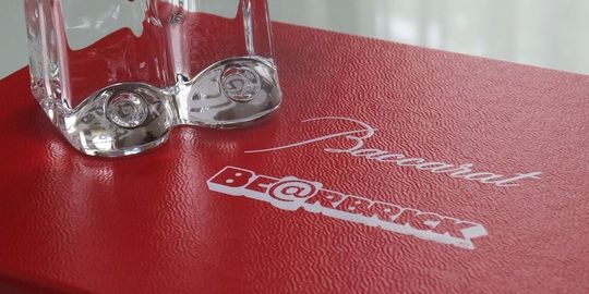 バカラのレッドボックスの上蓋に、『Baccarat』と『BE@RBRICK』と印刷されている写真。バカラの象徴的な赤い箱の蓋に、両ブランドのロゴが並んで印刷されており、この特別なコラボレーションを証明しています。