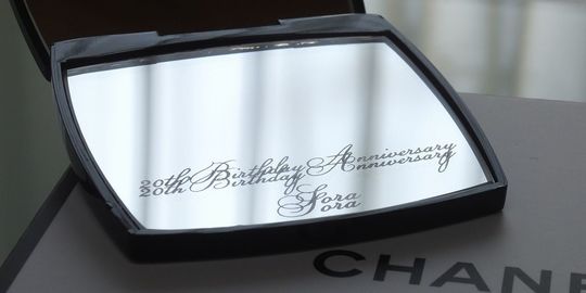 シャネルのミラー下部に、流麗なスクリプト体（筆記体）で『20th Birthday anniversary Sora』と名入れ彫刻を施した写真。二十歳の成人という人生の節目を祝うメッセージと名前が刻まれた、華やかでメモリアルなオーダーメイドギフトのイメージ。