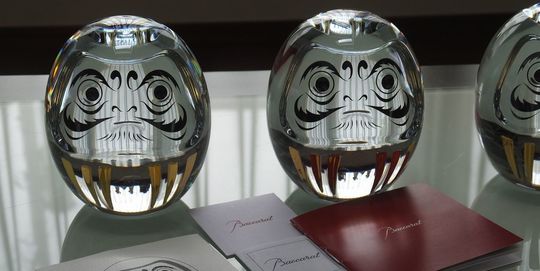 バカラのダルマ3体が、テーブルの上に置いてある写真。透明なクリスタルで象られた3体のダルマが、光を反射して輝き、日本の伝統的な縁起物とフランスのクリスタル技術が融合したユニークな魅力を放っています。名入れギフトとしても、特別な願いを込めるお守りとしても最適な贈り物です。