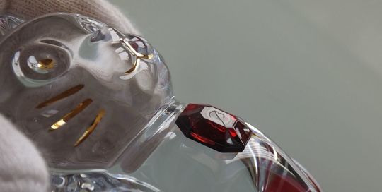 バカラの招き猫 レッドオクトゴンの胸元のレッドオクトゴンに『B』の刻印が入っているところからの横顔を映した写真。クリアクリスタル製の招き猫が、その横顔と共に、胸元の赤いオクトゴンに繊細に刻まれたバカラの『B』のロゴが光を反射して輝いています。ブランドの証と、幸運を招く縁起の良い置物としての高級感を際立たせています。