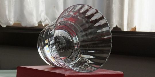 バカラの『ミルニュイ タンブラー』が、バカラのシンボルカラーである赤い純正ボックスの上に、横に倒して置かれている様子の写真。アラビアンナイトをイメージしたミルニュイ特有の優雅なプリーツカットと、深紅のボックスの強烈な色彩のコントラストが際立っている。タンブラーが横向きに置かれることで、そのクリスタルの複雑なカットと輝きがより強調され、最高級ギフトとしての存在感と美しさを視覚的に伝えている。