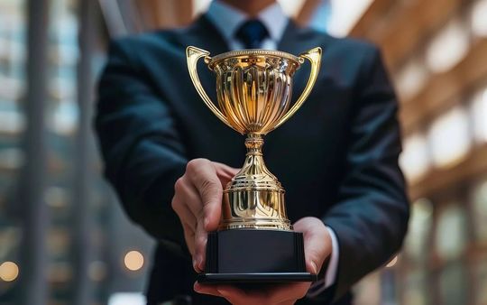 永年勤続表彰・サービスアワードの法人ギフト スーツ姿の男性が、輝く金のカップトロフィーを差し出す授与シーン