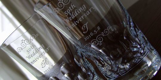 バカラのハイボールグラス、ベルーガハイボールグラスの側面に、『50th Wedding Anniversary T 2016.5.8』と、イニシャル『T』の部分が『H』と名入れ彫刻された写真。文字を挟むように装飾デザインが施されており、2客がテーブルの上に置かれています。重厚なクリスタルに、金婚式を祝う特別なメッセージと、繊細なデザインが美しく刻まれ、夫婦の絆を象徴する仙台からの特別な贈り物となっています。