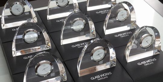 テーブルの上に、グラスワークスナルミのアーチクロックが大量に整然と並べられている写真。透明感のある光学クリスタルの時計が光を反射して輝き、企業の創立記念や永年勤続表彰などのために多数用意された記念品の壮観な様子を伝えるイメージ。